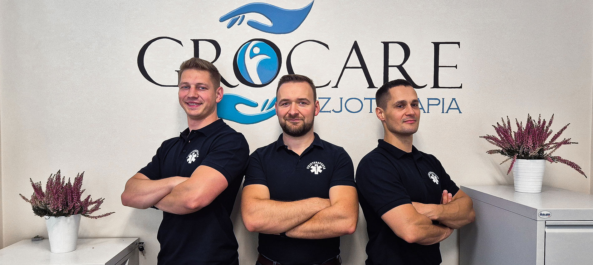 Zespół fizykoterapeutów Kamil Bojar, Grzegorz Grochal, Maciej Tobiasz na tle ściany z logiem Grocare Fizjoterapia